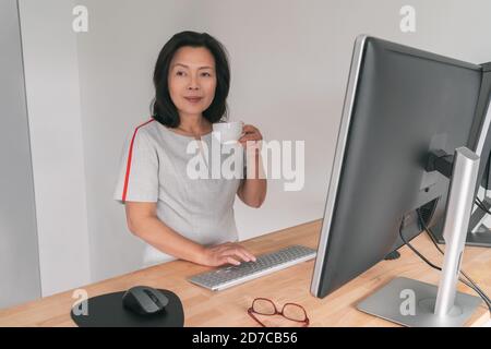 Donna d'affari asiatica matura che lavora sulla scrivania in piedi a casa ufficio bere caffè tazza prendere una pausa Businesswoman professionista signora al lavoro di digitazione Foto Stock