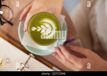 Vista dall'alto, bicchiere di tè verde matta caldo con art schiuma di tenuta da donna mano sul tavolo Foto Stock