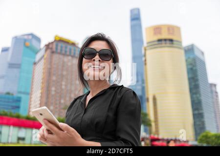 Asian business woman broker immobiliare utilizzando il telefono cellulare SMS a Shanghai città, Cina. Donna matura sorridente felice che lavora su smartphone all'aperto Foto Stock
