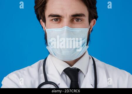 Medico in un cappotto bianco e maschera medica, con uno statoscopio intorno al collo vicino - su uno sfondo blu. Protezione concettuale contro virus e. Foto Stock