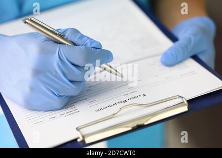 La mano con guanti tiene gli appunti e la penna e riempie il paziente modulo di registrazione Foto Stock