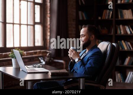 Caffè fresco al lavoro. Giovane sicuro in abbigliamento casual elegante che lavora su un computer portatile e beve una bevanda calda mentre si siede al lavoro Foto Stock