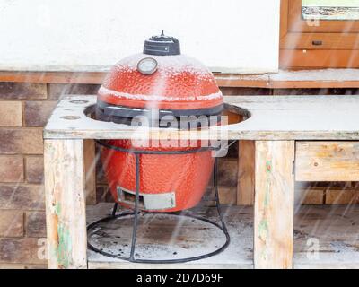 Barbecue rosso in ceramica si trova all'esterno nel foro di un tavolo in legno in una giornata invernale innevata Foto Stock