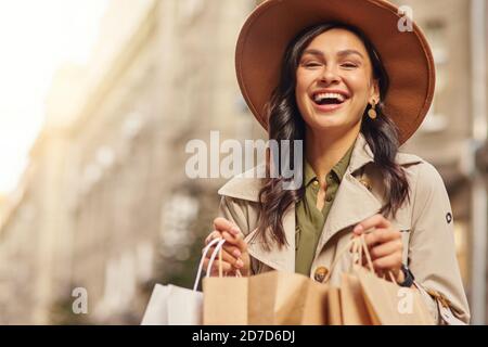 Shopping come terapia. Ritratto di una giovane donna eccitata che indossa cappotto grigio e cappello che tiene borse per lo shopping, in piedi sulla strada della città e sorridendo alla macchina fotografica. Moda, concetto di stile di vita della gente Foto Stock