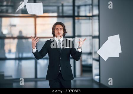 Cammina in ufficio e getta i documenti in aria. Ritratto di bel giovane uomo d'affari in abito nero e cravatta Foto Stock