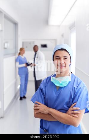 Ritratto di Dottore Maschile che indossa scrub in piedi in ospedale Busy Corridoio Foto Stock