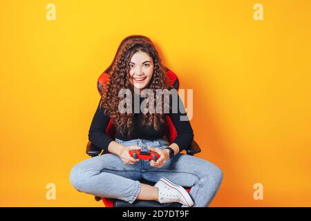 Donna seduta dietro la sedia da gioco nelle mani che tiene in rosso gamepad su sfondo giallo Foto Stock