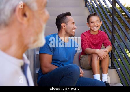 Sorridente Famiglia ispanica maschile Multi-Generation seduta sui gradini in Giardino E parlare insieme Foto Stock