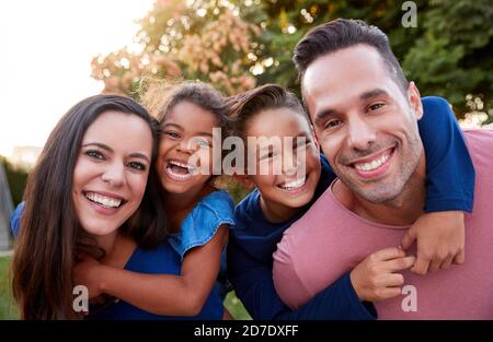 Ritratto di sorridente famiglia ispanica con genitori che danno figli Piggyback Passeggiate in giardino a casa Foto Stock