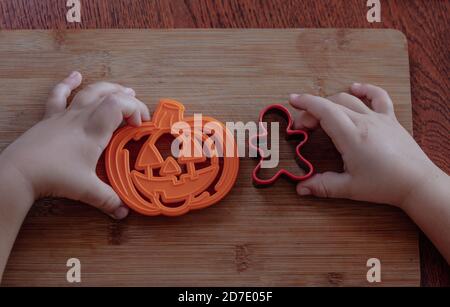 Le mani di piccolo capretto che fanno i biscotti tradizionali di Halloween. Taglierine per i biscotti di festa su un fondo di tavola di legno. Preparazione dei cookie. Fasi di Foto Stock