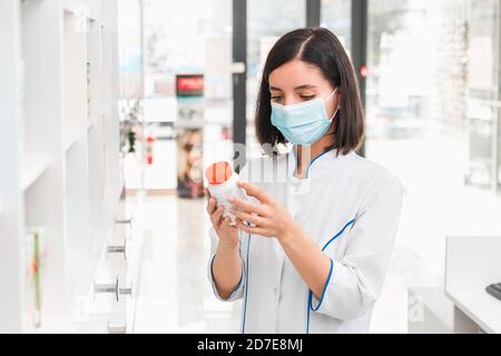 scegliere la medicina giusta. professionale cercando farmacista femminile con maschera medica in negozio di droga studiare il prospetto di un nuovo farmaco Foto Stock