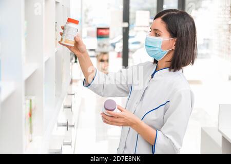 scegliere la medicina giusta. professionale cercando farmacista femminile con maschera medica in negozio di droga studiare il prospetto di un nuovo farmaco Foto Stock