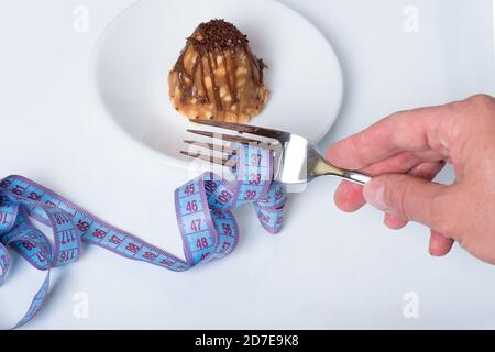 Una mano femmina tiene una forchetta con un nastro di misurazione a spirale, da dietro una torta su un piatto su sfondo bianco, primo piano, spazio di copia. Dieta e peso lo Foto Stock