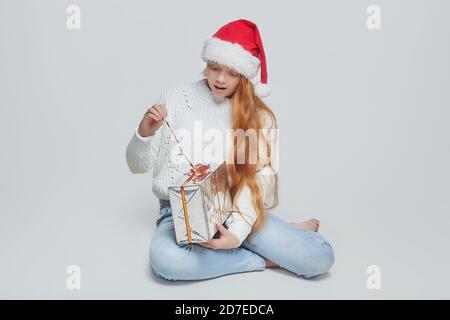 Una ragazza dai capelli rossi che indossa un cappello Babbo Natale apre un regalo. Una sessione fotografica in studio su sfondo bianco Foto Stock