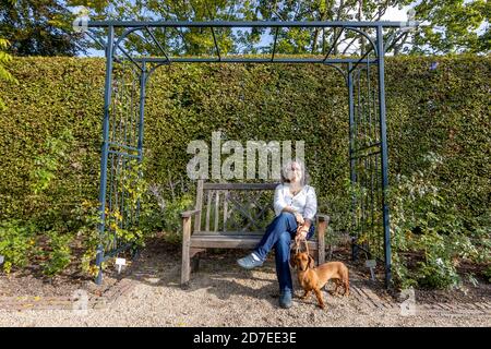 Sorridente donna messicana matura indossando occhiali seduti su una panca di legno, il suo dachshund in piedi accanto a lei con vegetazione verde sullo sfondo, it Foto Stock