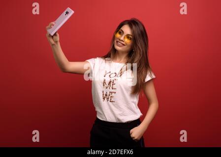 Ritratto di bella bella donna felice facendo selfie isolato su sfondo rosso, indossando T-shirt bianca e occhiali gialli. Concetto di tecnologia Foto Stock