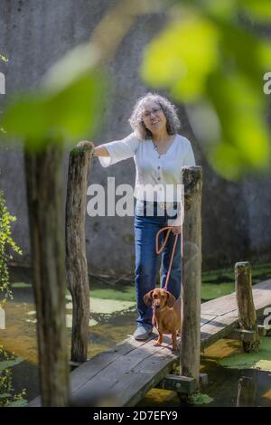 Sorridente donna messicana matura con bicchieri in piedi accanto al suo dachshund su un percorso di legno sopra uno stagno con una parete rocciosa sullo sfondo, godendo di un Foto Stock