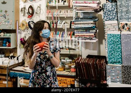 Giovane ragazza con lunghi capelli marroni che indossa una maschera facciale e sorridente con una palla di lana rossa in un haberdashery. Abito con dettagli floreali. Naturalezza Foto Stock