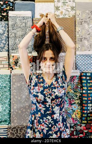 Ritratto di una giovane ragazza che tiene i suoi lunghi capelli su uno sfondo di tessuti di vari colori. Abito con dettagli floreali. Foto Stock