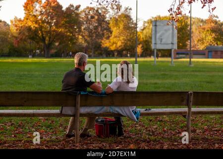 Una coppia caucasica di mezza età è seduta su una panchina di legno in un parco coperto di foglie cadute al tramonto. Un concetto d'autunno scenico con romanticismo e t Foto Stock