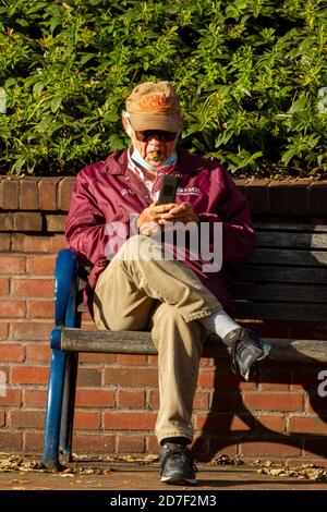 Frederick, MD, USA 10/14/2020: Un uomo caucasico anziano che indossa una maschera è seduto a croce zampe su una panchina del parco con un sigaro in bocca, cercando Foto Stock