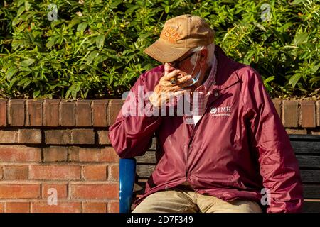 Frederick, MD, USA 10/14/2020: Un uomo caucasico anziano è seduto da solo su una panchina di strada e fuma un sigaro spesso. Mette la sua maschera contro CO Foto Stock