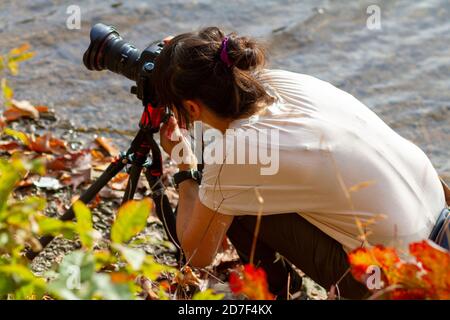 Una giovane donna caucasica che indossa abiti casual è inginocchiata a terra da un fiume. Sta cercando di comporre una fotografia di paesaggio con la sua dslr ca Foto Stock