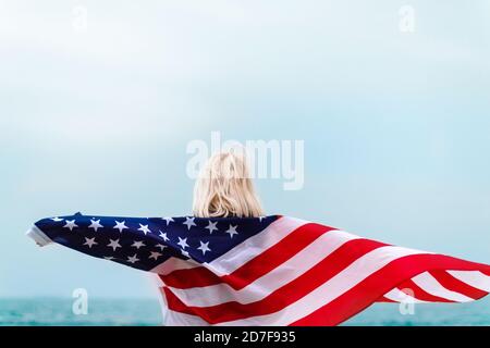 Donna bionda caucasica che tiene la bandiera americana appoggiata contro la schiena e. guardando al mare Foto Stock