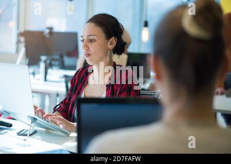 Colleghi multietnici biondi caucasici e Latina capelli scuri belle donne d'affari (dipendente) seduto su un divano giallo mentre si lavora su un portatile dentro Foto Stock