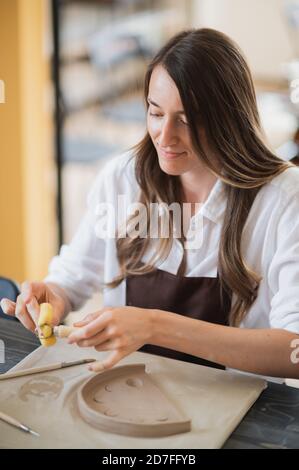 Donna che lavora con l'argilla con le mani e l'utensile da taglio ad anello da vicino in un laboratorio di ceramica. Foto Stock