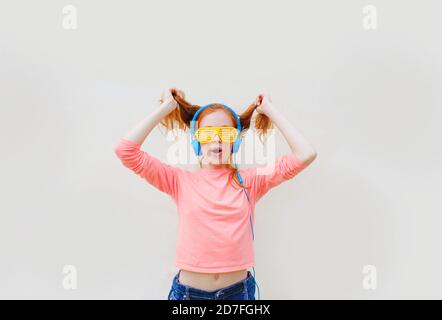 una ragazza adolescente con capelli rossi che indossa occhiali gialli e. le cuffie blu ballano vicino a una parete bianca Foto Stock