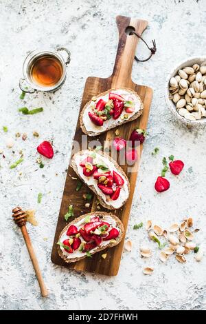 Sandwich con fragole, formaggio morbido, pistacchi, menta e miele su tavola di legno su sfondo grigio. Vista dall'alto. Foto Stock