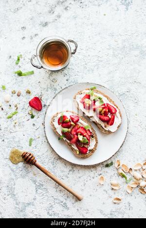 Sandwich con fragole, formaggio morbido, pistacchi, menta e miele su tavola di legno su sfondo grigio. Vista dall'alto. Foto Stock