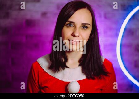 Giovane donna in tuta di Babbo Natale sorridente sullo sfondo di una parete illuminata. Ritratto di bella brunetta in costume di Natale. Concetto di Natale Foto Stock