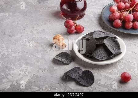 Patatine nere con carbone, aceto balsamico in vetro, uva rossa su un piatto di ceramica blu su fondo grigio di cemento. Vista laterale, spazio copia. Foto Stock