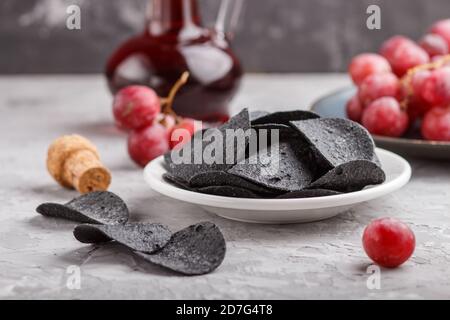 Patatine nere con carbone, aceto balsamico in vetro, uva rossa su un piatto di ceramica blu su fondo grigio. Vista laterale, primo piano, se Foto Stock