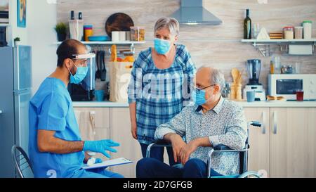 Medico durante la visita a casa utilizzando un termometro a infrarossi su un uomo anziano in sedia a rotelle disabili per controllare la temperatura corporea. Persone anziane disabili che ricevono un consiglio da un lavoratore sociale Foto Stock