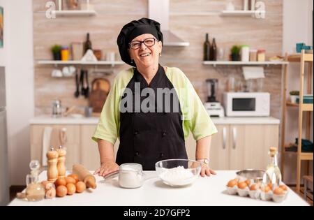 Donna anziana felice che indossa bonete guardando la macchina fotografica nella sala da pranzo a casa. Panettiere anziano in pensione in cucina uniforme preparare ingredienti di pasticceria su tavola di legno pronto a cucinare pane gustoso fatto in casa. Foto Stock