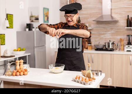 Panettiere preparazione di impasto cracking uova su farina di grano in cucina casalinga. Cuoco di pasta anziano che coglie l'uovo sulla ciotola di vetro per la ricetta della torta in cucina, mescolando a mano, impastando. Foto Stock