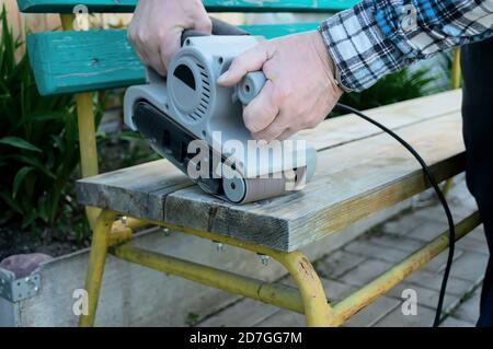 Abbellimento del giardino. Handyman affila una panca di legno sulla strada nel giardino. Master rimuove uno strato di vernice asciutta dalla superficie di legno Foto Stock