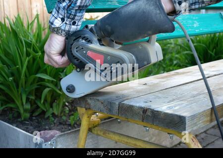 Abbellimento del giardino. Handyman affila una panca di legno sulla strada nel giardino. Master rimuove uno strato di vernice asciutta dalla superficie di legno Foto Stock