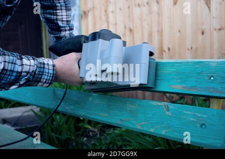 Abbellimento del giardino. Handyman affila una panca di legno sulla strada nel giardino. Master rimuove uno strato di vernice asciutta dalla superficie di legno Foto Stock