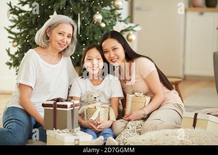 Ritratto di felice madre asiatica con le sue due figlie sorridenti Alla macchina fotografica che si siede sul pavimento celebrano insieme il Natale Foto Stock