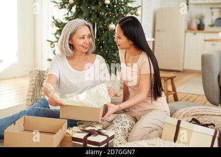 Nipote che apre il regalo di Natale insieme a sua nonna loro Festeggiamo il Natale a casa Foto Stock