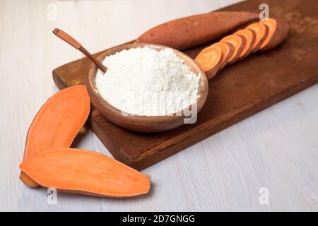 farina di patate dolci in un piatto di legno su sfondo chiaro. patate dolci fresche tagliate a pezzi d'arancia accanto alla farina. vista dall'alto. clo Foto Stock