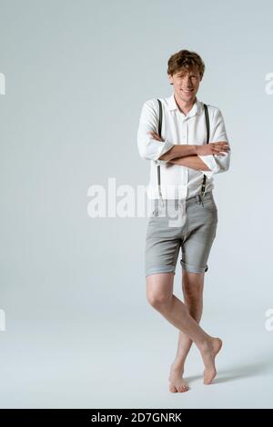 L'uomo elegante in sospetti ha attraversato le braccia in piedi nudi in piena altezza in studio su sfondo bianco. Affascinante dude Foto Stock