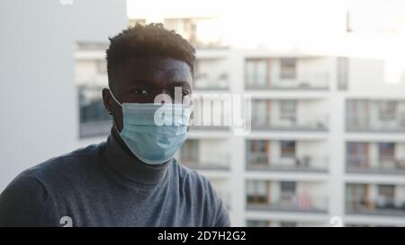 Uomo nero con maschera medica sul balcone che guarda alla macchina fotografica. Problemi di quarantena e di salute mentale. Foto di alta qualità Foto Stock