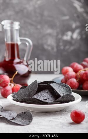 Patatine nere con carbone, aceto balsamico in vetro, uva rossa su un piatto di ceramica blu su fondo grigio. Vista laterale, primo piano, se Foto Stock