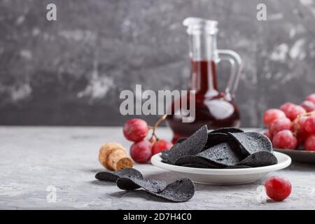 Patatine nere con carbone, aceto balsamico in vetro, uva rossa su un piatto di ceramica blu su fondo grigio. Vista laterale, primo piano, se Foto Stock