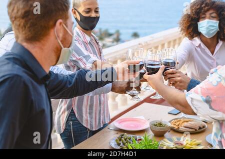 Giovani amici multirazziali che tostano con bicchieri di vino rosso mentre indossano maschera protettiva per il viso durante la pandemia del virus corona Foto Stock